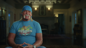 A smiling older man with tan light-toned skin and a bushy blond mustache/fu manchu, sitting in a nice living room-esque setting.