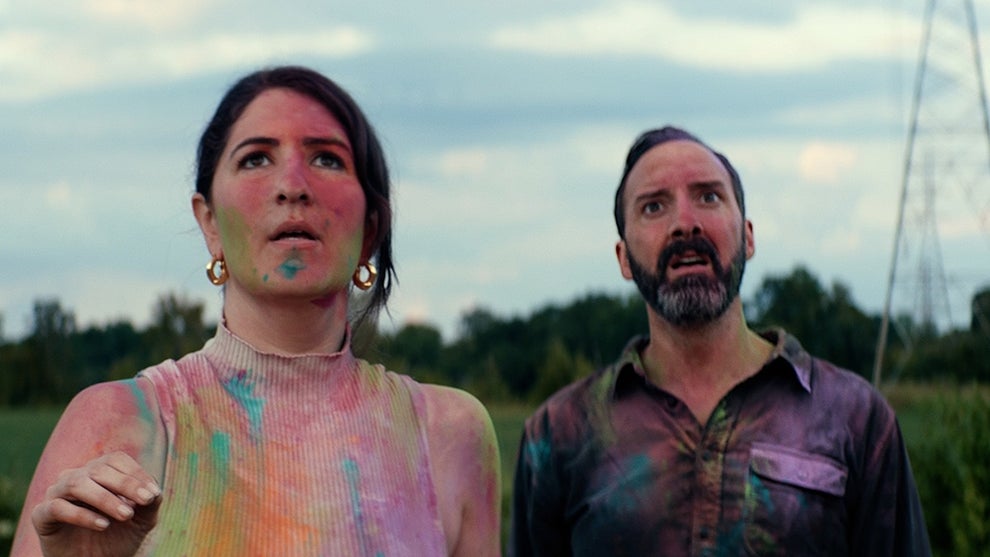 A woman and a man with their faces and clothes covered in various bright paints stand outside in a green field, a power line tower and various trees in the background.