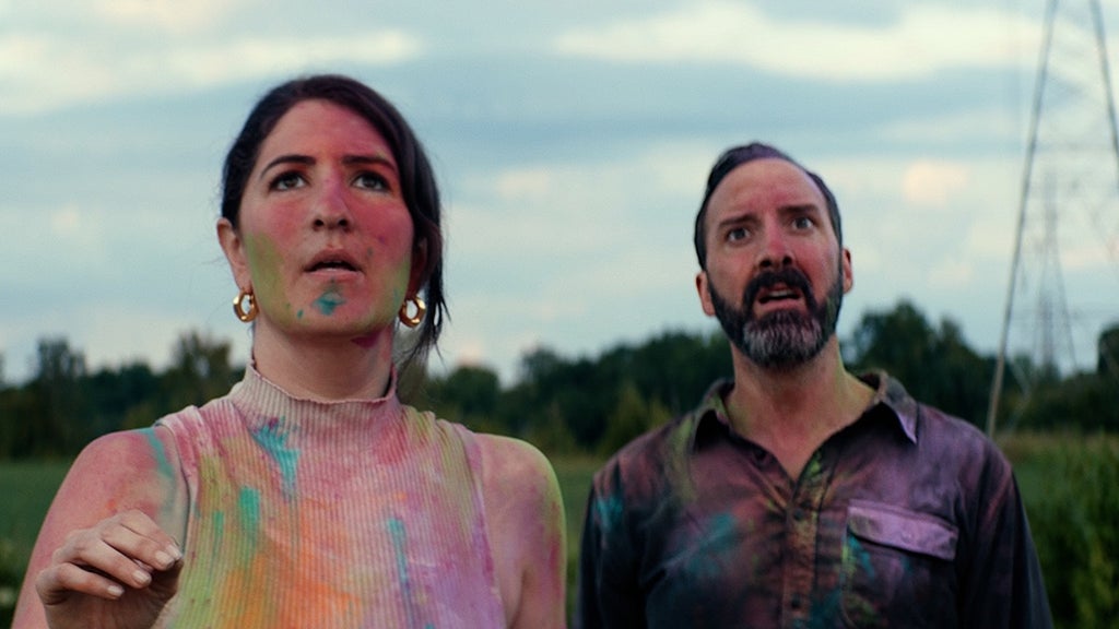 A woman and a man with their faces and clothes covered in various bright paints stand outside in a green field, a power line tower and various trees in the background.