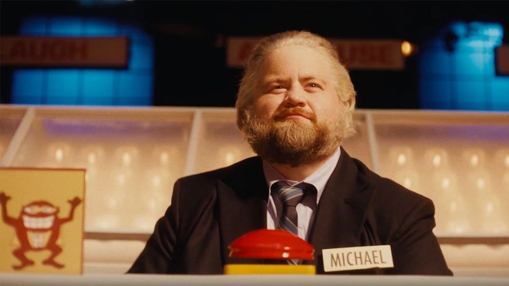 A man with graying hair and a darker beard shows a confident look as he sits on a game show set with a nametag reading "Michael," a red buzzer button in front of him — and a strange looking creature in a drawing on the left.