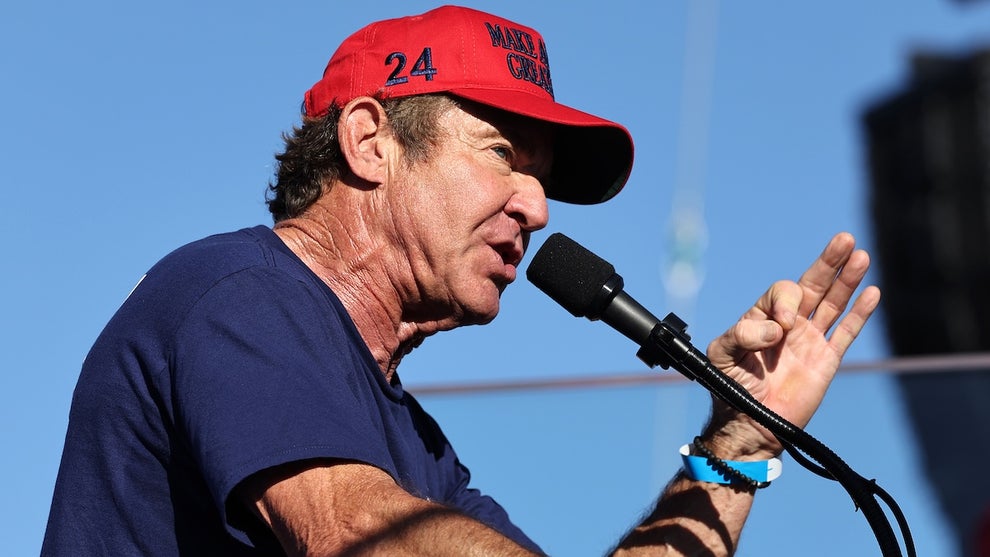 An older man wears a red hat and makes an emphatic gesture as he speaks in front of a microphone outdoors.