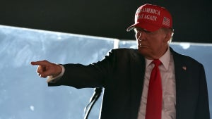 A man with light-toned skin, wearing a suit and a red baseball cap reading "MAKE AMERICA GREAT AGAIN," points toward a crowd as he stands behind bulletproof glass.