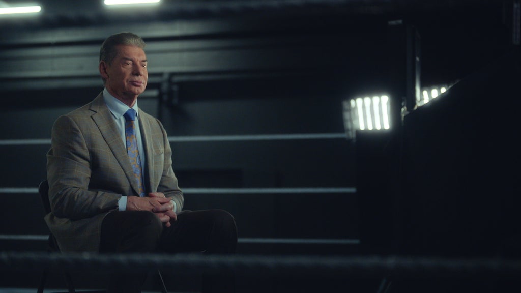 An older man wearing a suit sits in a chair in a wrestling ring. He has light-toned skin.