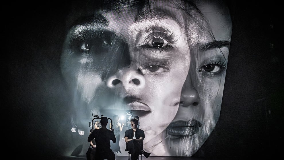 Two people sit on stage while giant projected images of their faces loom over behind them.
