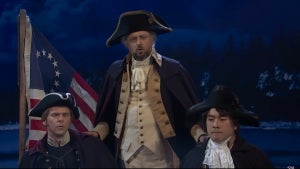 A group of three men in a boat, with an old American flag with just 13 stars behind them and dim twilight lighting. Two of the men sit in the boat and one stands. The man standing and the man on the left have light-toned skin, while the man to the right has medium-toned skin.