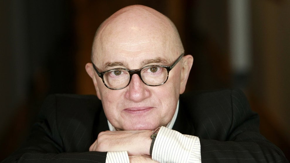 An older man with light-toned skin smiles and poses for the camera, wearing a suit.