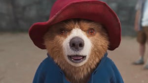An image of a CGI bear with an open mouthed expression, wearing a red hat and blue coat, in a jungle setting.