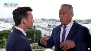 An older man animatedly gestures while speaking with a younger man outside near a body of water, both wearing suits. They each have light-toned skin.