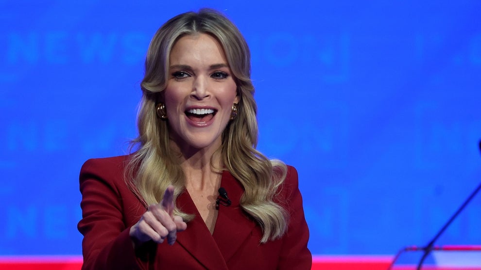 A woman in a red suit smiles as she stands on stage. She has light-toned skin.