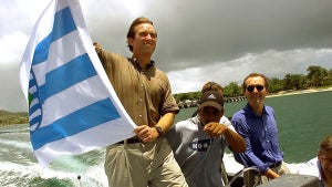 A man with light-toned skin waves a flag in a small speedboat, with others near him with medium-toned skin.