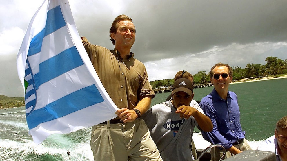 A man with light-toned skin waves a flag in a small speedboat, with others near him with medium-toned skin.