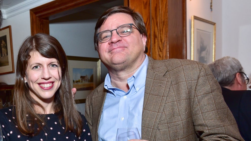 A woman and a man with light-toned skin pose for a photo at a party in a home. The man holds wine.