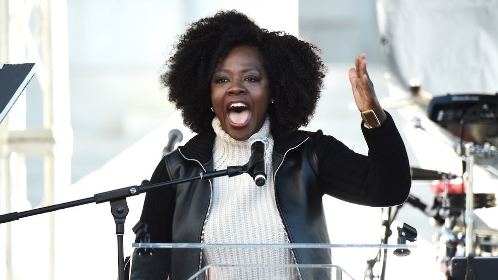 A woman makes an animated gesture standing at a podium. She has dark-toned skin and speaks into a microphone.