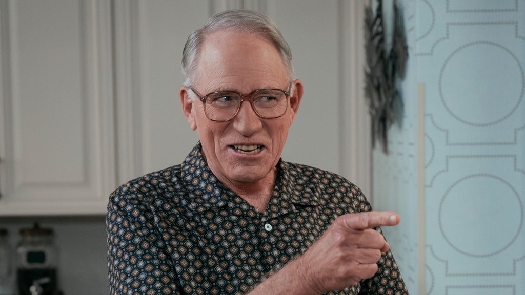 An older man wearing glasses smiles and points his finger at someone