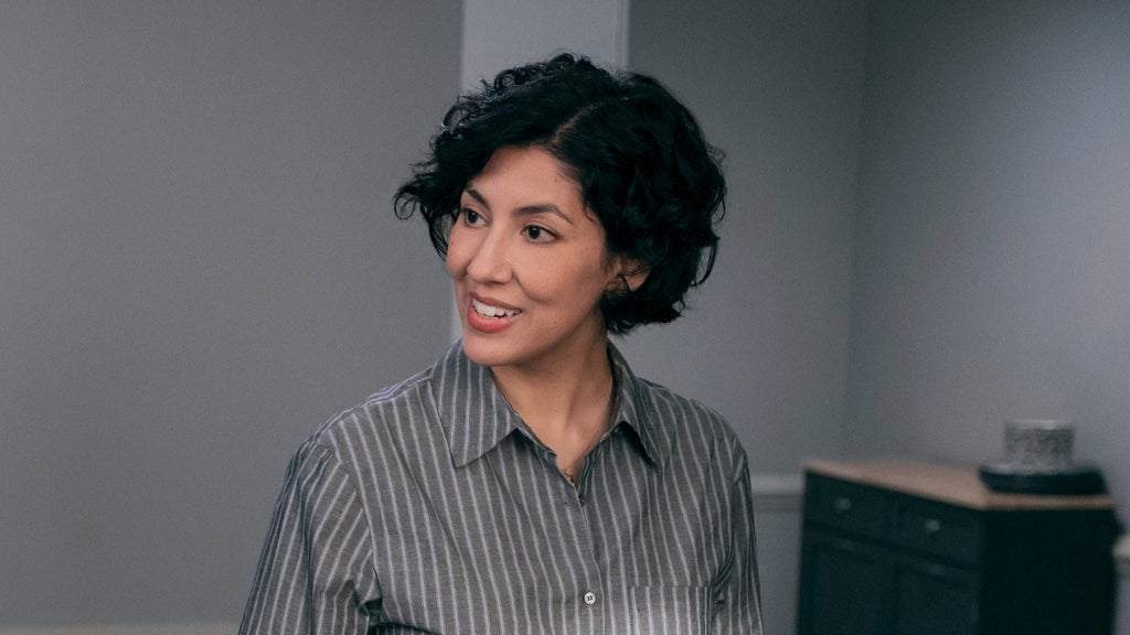 A woman with short brown hair and a striped collared shirt smiles at someone standing to her left