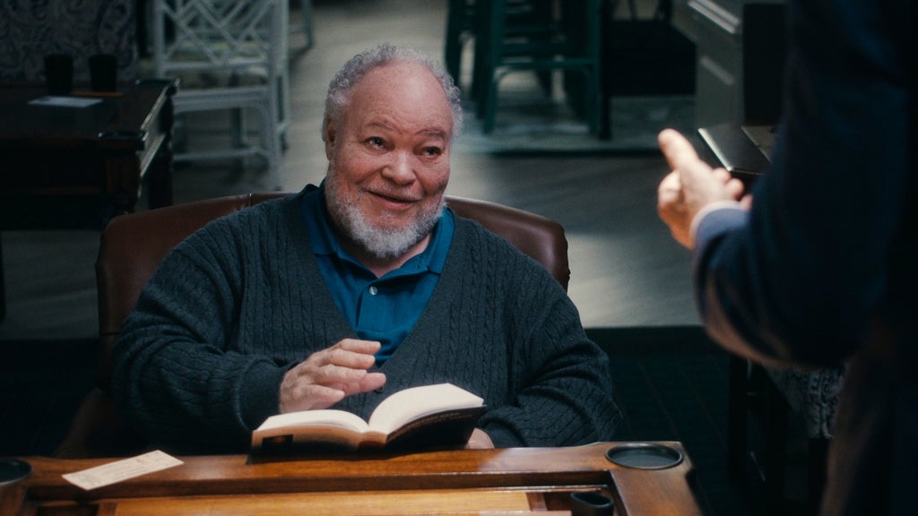 An older man with grey hair and a beard smiles while holding a book