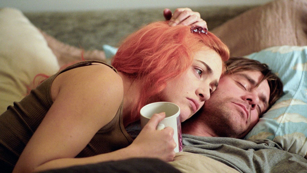 A woman with orange hair lies on a bed, propped up on her side, holding a white mug, propped up on a man's chest. She looks down at a man with short brown hair who is resting his head on a pillow, his eyes closed. The bed has light-colored sheets and pillows, and the room has soft, natural lighting.