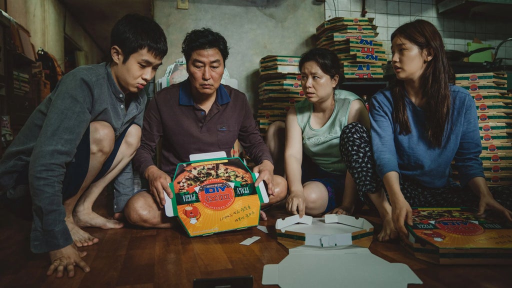 Four people are crouched around a wooden table, intently looking at several pizza boxes. The boxes have colorful designs with text, including one prominently displaying the word PIZZA. Behind them, stacks of flat pizza boxes are piled against the wall. The setting appears to be a small, cluttered room with tiled walls and various items in the background. The group seems focused and engaged with the contents of the boxes.