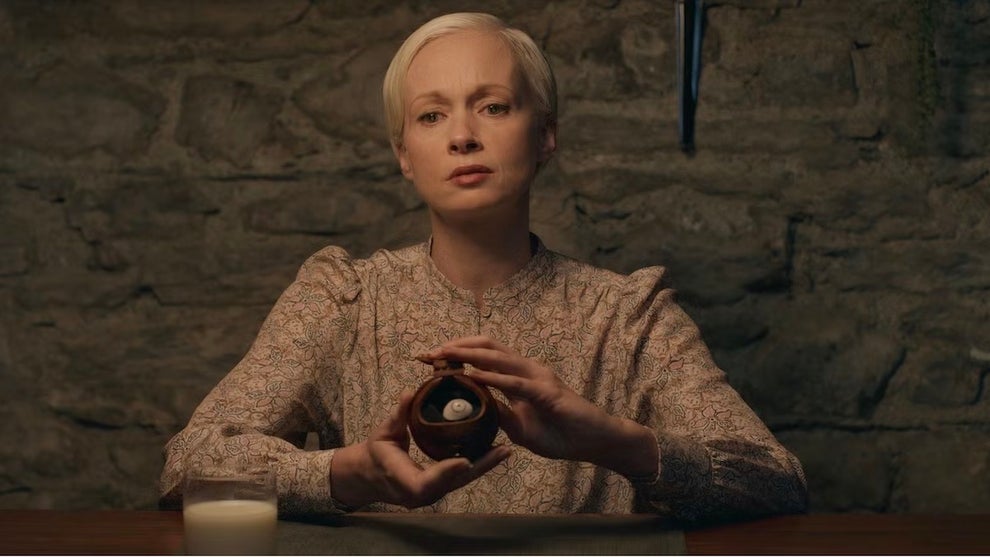 A woman with short blonde hair sits at a wooden table in a dimly lit stone room. She wears a patterned beige dress with puffed sleeves. In her hands, she holds a small orb with a prosthetic eye inside. A candle is on the table to her left. The background shows a rough stone wall.