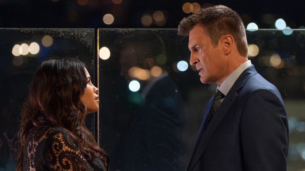 Two individuals are engaged in a conversation in a dimly lit setting with blurred lights in the background, suggesting a nighttime scene. One person is wearing a dark patterned outfit with long hair, facing the other person who is dressed in a formal suit with a tie, and has short hair. They are standing in front of a glass panel, which reflects some of the surrounding lights. The atmosphere appears to be serious or contemplative.