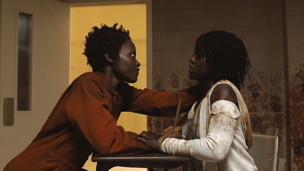 Two individuals who look alike are seated closely together indoors, facing each other. One person, wearing an orange shirt, is holding a pair of scissors and appears to be pointing them at the other person in a threatening manner, who is dressed in a white long-sleeve top. The setting is dimly lit with warm, yellowish lighting coming from a window in the background. The room has plain walls and a simple, minimalistic decor.