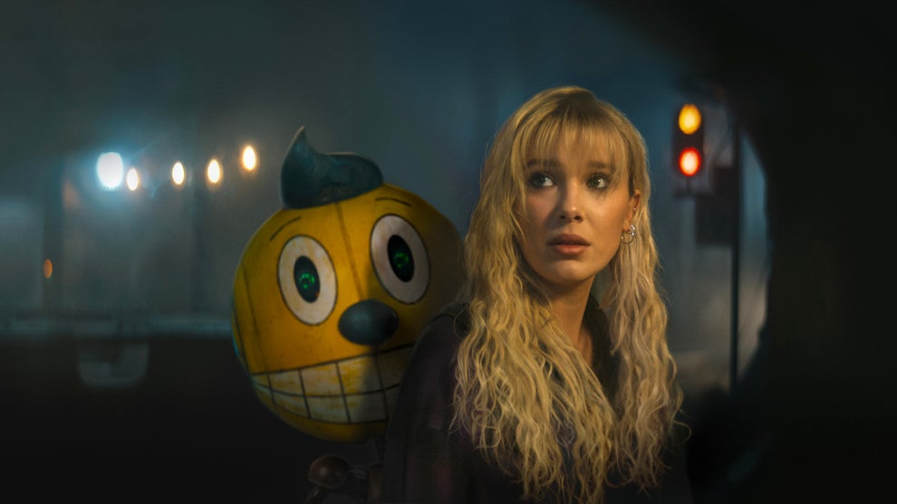 A young woman with blonde hair and bangs stands beside an animated robot character resembling a yellow jack-o’-lantern with a smiling face and glowing green eyes. The woman is wearing a dark outfit and has dangling earrings. The background is dimly lit with blurred, colorful lights, creating a mysterious atmosphere. The setting appears to be indoors with a hint of fog.