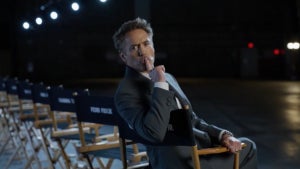 A man in a gray suit sits in a director’s chair on a dimly lit stage, his hand resting on his chin in a thoughtful pose. Behind him, rows of empty chairs are visible, labeled in white text. The background is dark, with stage lights illuminating the scene.