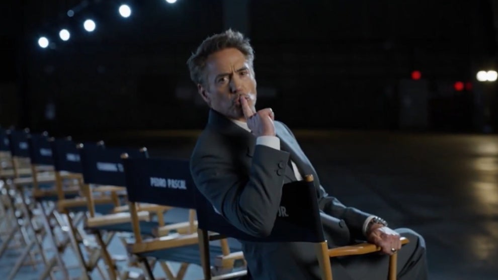 A man in a gray suit sits in a director’s chair on a dimly lit stage, his hand resting on his chin in a thoughtful pose. Behind him, rows of empty chairs are visible, labeled in white text. The background is dark, with stage lights illuminating the scene.