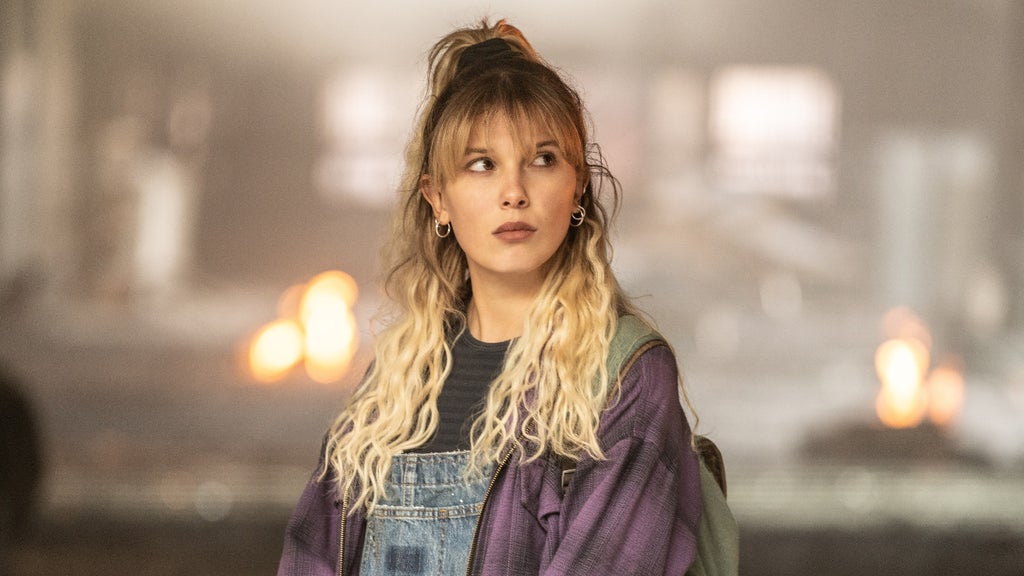A young woman with blonde hair stands in a softly lit room, looking off to the side with a contemplative expression. She is wearing a dark top, denim overalls, and a purple plaid shirt draped over her shoulders. Her hair is styled with a loose bun on top, and she has on dangling earrings. The background is blurred with warm, glowing lights creating a bokeh effect.
