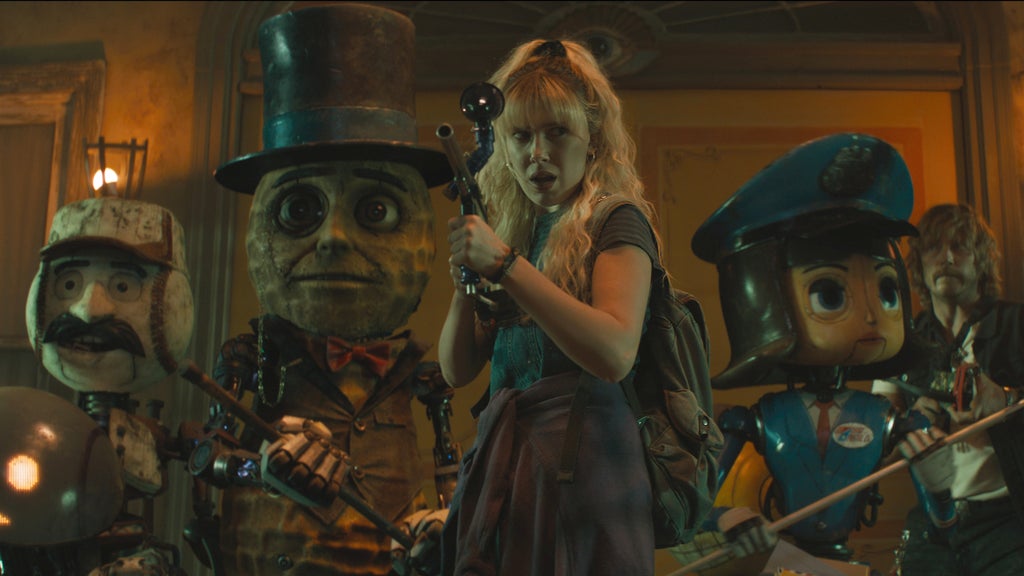 A young woman with blonde hair stands in the center of a dimly lit room, holding a flashlight. She wears a dark dress and carries a backpack. Surrounding her are several animated characters resembling old-fashioned dolls or puppets, each with exaggerated features and costumes, including a top hat, a beret, and various accessories. The setting appears to be indoors with warm lighting and wooden details.