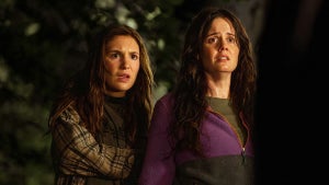 Two women stand side by side outdoors at night, looking up with expressions of concern or surprise. One wears a plaid shirt over a black top, while the other wears a purple sweater and has a distressed look on her face. The background is dark with foliage visible.