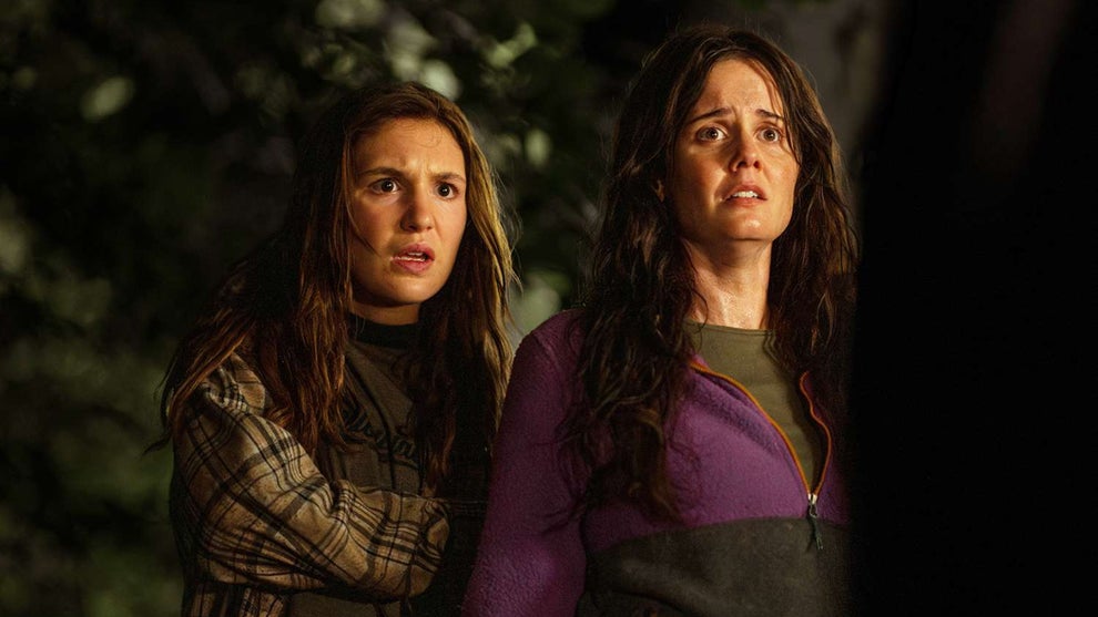 Two women stand side by side outdoors at night, looking up with expressions of concern or surprise. One wears a plaid shirt over a black top, while the other wears a purple sweater and has a distressed look on her face. The background is dark with foliage visible.