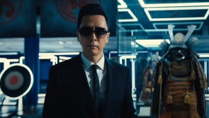 A man in a dark suit, white shirt, and tie, wearing sunglasses, stands in a futuristic room with blue and white lighting. Behind him is a traditional Japanese samurai armor display and a circular target with a bullseye. The environment looks clean, polished and high-tech.