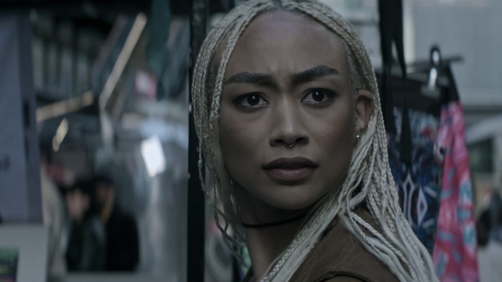 A woman with long, platinum blonde braids and a septum piercing stands in a bustling outdoor market, looking off to the side with a tense, suspicious expression. Her brows are furrowed, and her gaze is focused, as if she’s just spotted something alarming or unexpected.