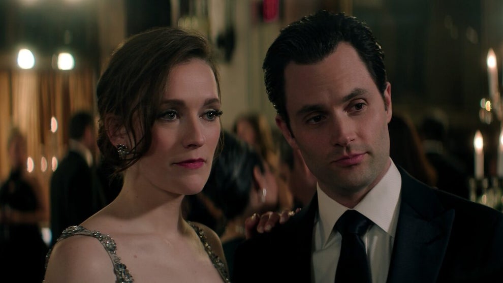 A well-dressed couple stands closely together at a formal event, surrounded by other elegantly dressed guests. The woman wears a sparkling, silver evening gown with delicate straps, her expression composed but slightly tense. The man beside her, in a black suit and tie, has a confident, unreadable look on his face. Warm lighting and soft-focus candles in the background create an upscale, moody atmosphere that suggests secrets may be hiding behind the glamour.