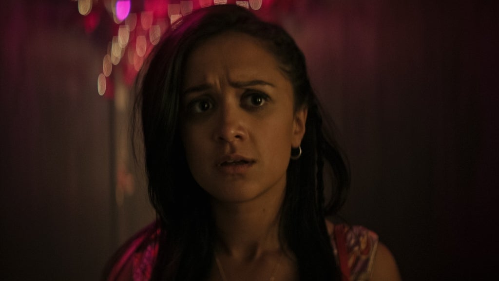 A young woman with long dark hair and hoop earrings stands in a dimly lit hallway illuminated by soft pink and purple lights. Her expression is tense and alarmed, eyes wide and brows furrowed, as if she’s just seen or heard something unsettling. The background is blurred, enhancing the focus on her emotional reaction.