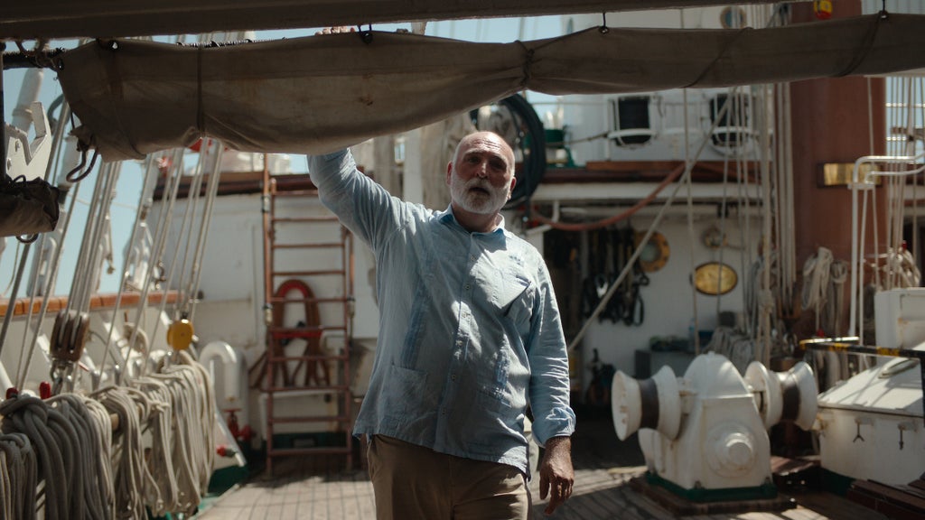 A man with a gray beard and light blue shirt stands on the deck of a ship, holding onto a rolled-up sail above him. He is surrounded by ropes, nautical equipment, and wooden features of the boat, bathed in daylight.