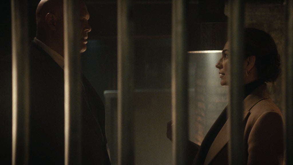 A tall bald man in a black suit and a brunette woman in a brown suitcoat are seen lookin at each other intently and seriously through the bars of a jail cell.