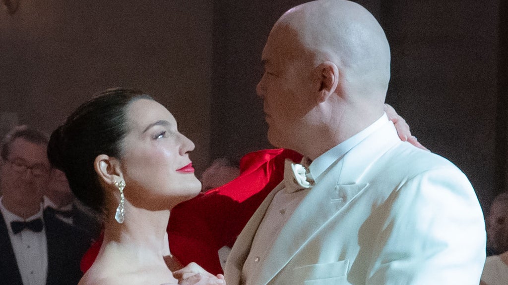 A brunette woman with an updo, wearing red lipstick, a red dress and glimmering jewel earings, dances with a tall bald man in an all white suit. They stare into each other's eyes.