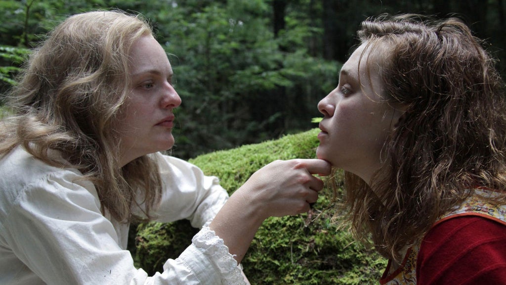 Two individuals, one with light hair and wearing a white shirt, and the other with curly hair and wearing a red top, are in a lush, green outdoor setting. The person in the white shirt is gently touching the chin of the person in the red top, who is looking up at them. The background features dense greenery and trees, suggesting a forest or garden environment.