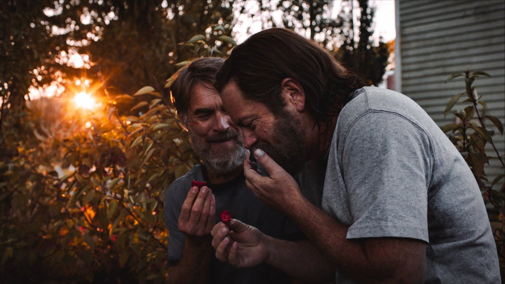 Two men are outdoors, one with a beard and glasses wearing a dark shirt, the other with longer hair wearing a gray shirt. The bearded man is holding a strawberry and smiling, while the other man leans in, also smiling, holding a strawberry and covering his face while laughing. Behind them is lush greenery and a blurred, sunlit background.