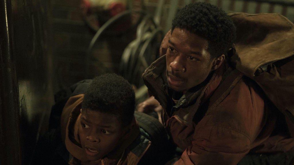 Two people crouch indoors in a dimly lit room, peering cautiously from behind a wooden structure. The person on the left wears a hooded jacket and has short, curly hair, while the person on the right has a serious expression, curly hair, and wears a brown jacket over a dark shirt. The background is shadowy with hints of red lighting.