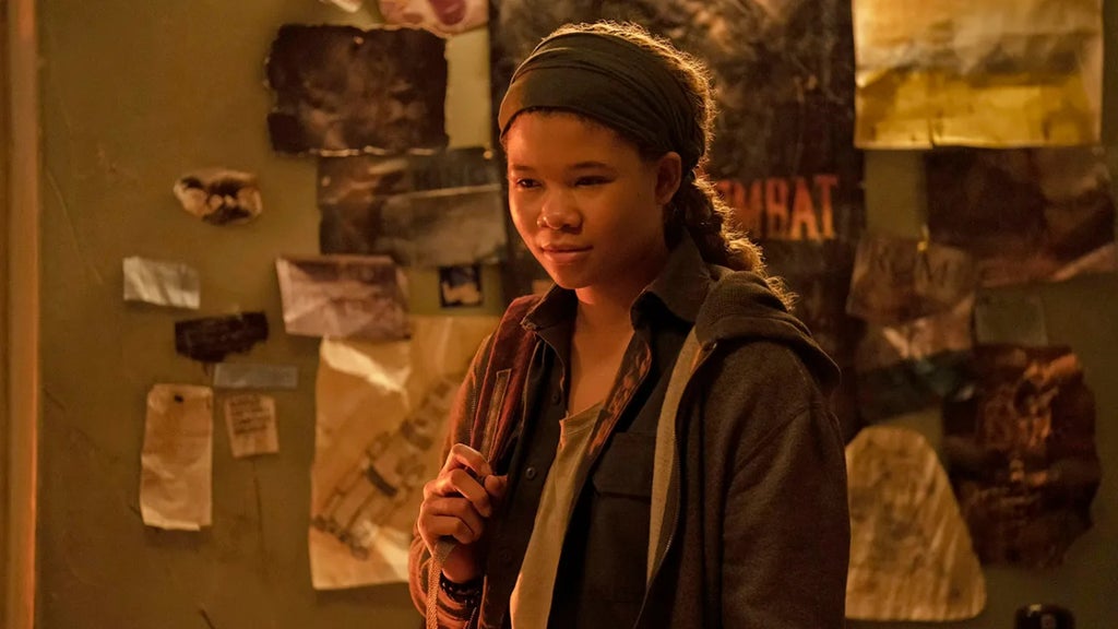 A person with a headband stands indoors, holding the strap of their backpack. They wear a dark jacket over a light shirt. Behind them, a wall is covered with various papers, images, and notes, partially obscured by shadows. The room is dimly lit with warm, golden light.