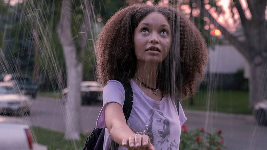 A person with curly hair stands outdoors near a spraying fountain, looking upward with a thoughtful expression. They wear a light purple t-shirt with a graphic design, a necklace, and a black backpack with a strap across their chest. The background shows a grassy suburban area with parked cars and trees.