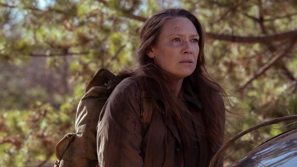 A woman with long brown hair stands outdoors near a car, looking upward with a serious expression and scratches on her face. She wears a dark jacket and a large brown backpack. Behind her, the setting is a wooded area with trees and foliage, and sunlight filters through the branches.
