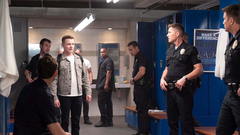 A tense scene in a police locker room shows several officers, some in uniform and others in casual clothes, gathered around a young man wearing a light gray jacket and backpack. Blue lockers are in the background and motivational posters are on the lockers.