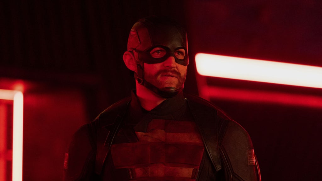 A man in a dark superhero suit and helmet stands in a room lit by red neon lights. His outfit features a stylized star and American flag patches, and his face is partially visible through a black mask. He has a short beard and a serious expression