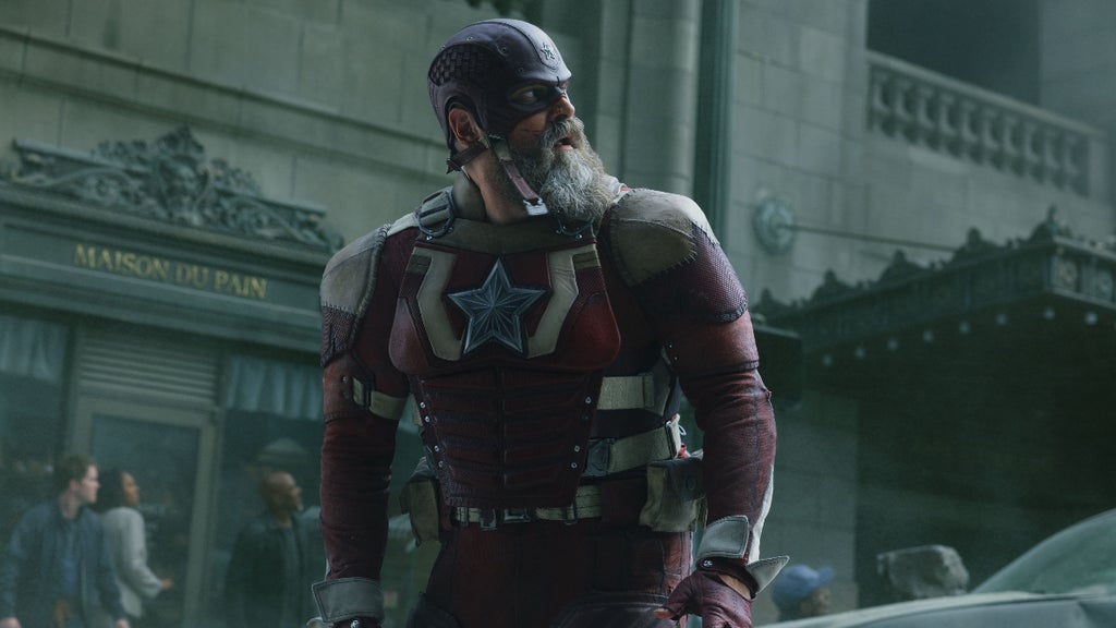 A muscular man in a red and gray armored suit stands alert in an urban setting, wearing a helmet with a star emblem and a full gray beard. His uniform, marked by a large star on the chest and utility belts at his waist, resembles a patriotic-themed super-soldier.