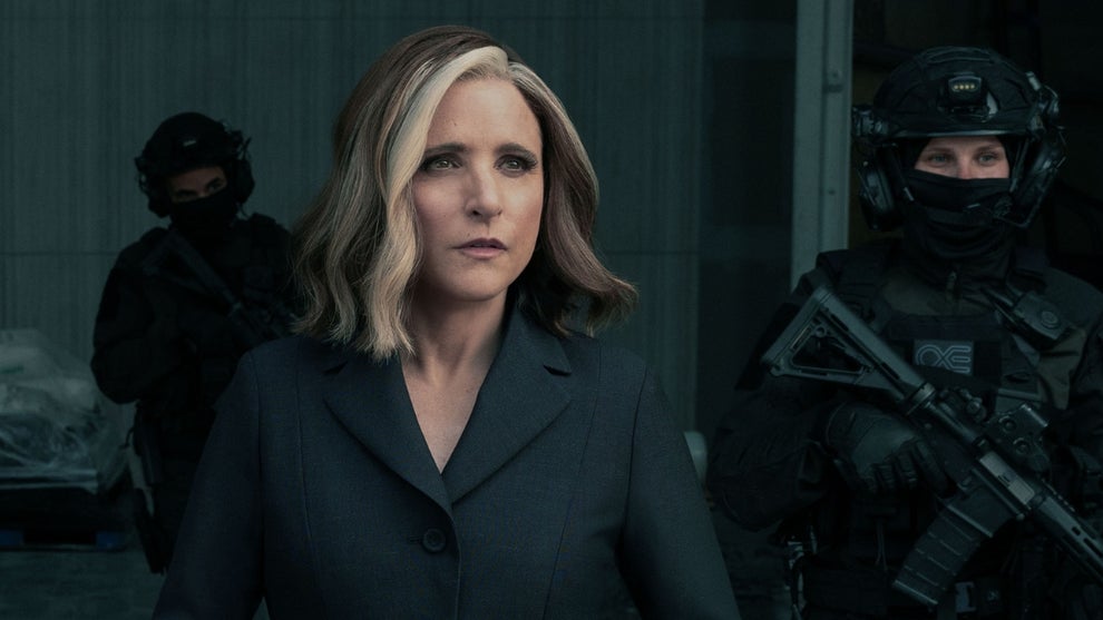 A woman in a dark suit stands in the foreground with a serious expression, looking slightly off-camera. Behind her, two individuals in tactical gear and helmets are positioned, one holding a futuristic rifle. The setting appears to be a dimly lit, industrial or military-style environment.