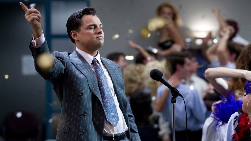 A man in a pinstripe suit, white dress shirt, and patterned tie stands speaking into a microphone. He points upward with his right hand. Behind him, blurred figures are raising their hands, while gold confetti falls through the air. The setting appears to be indoors with a blue-gray background.